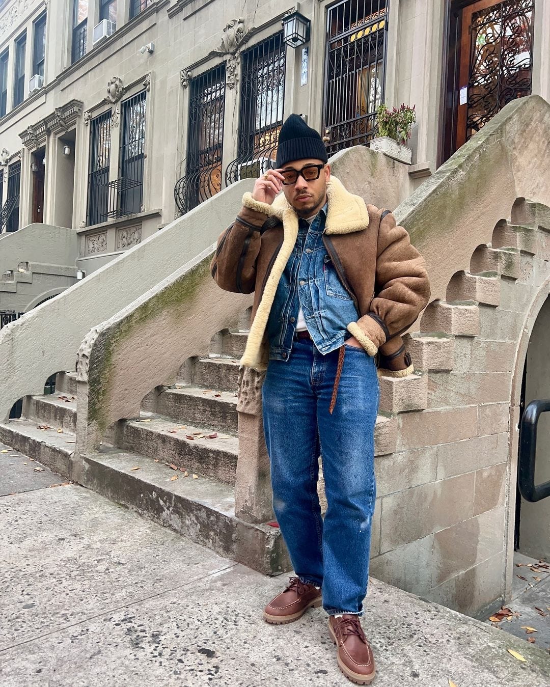 stylish young man wearing an oversized bomber jacket with sherpa collar over a denim jacket with blue jeans, a braided brown leather belt, and brown lace-up shoes