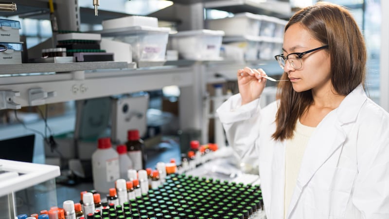 A woman in a white lab coat smells a scent sample in a lab filled with chemistry equipment.