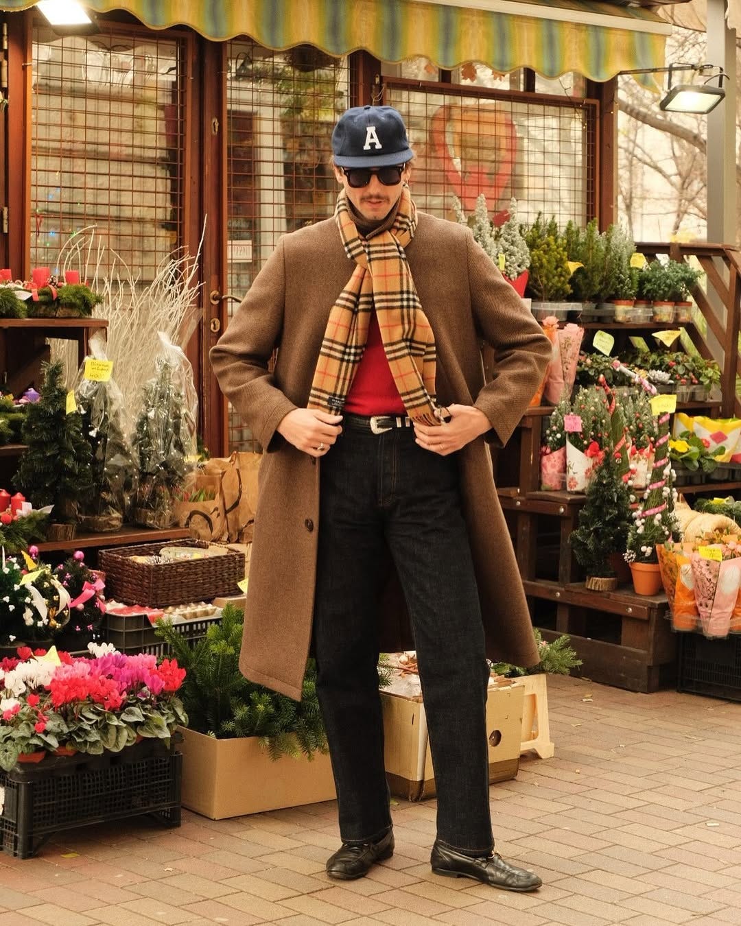 stylish man standing outside in front of a stand of flowers in a men's holiday party outfit, wearing a long brown wool topcoat with a plaid burberry scarf over a red sweater and black jeans with black loafers