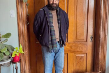 stylish man standing in front of a wooden door wearing a waist-length coat with a plaid wool scarf, sweater, jeans, and a beanie