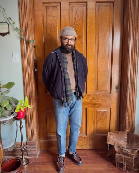 stylish man standing in front of a wooden door wearing a waist-length coat with a plaid wool scarf, sweater, jeans, and a beanie