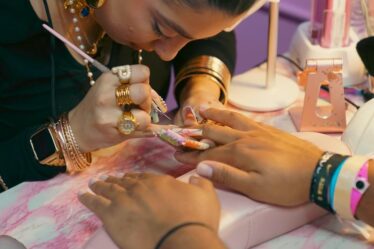 A woman wearing various wristbrands for different events is getting her nails done. The woman doing her nails wears lavishly long acrylics in Floridian sunset tones, some branded with the word NIKE or the brand's logo. She also wears a stack of gold bracelets and several chunky rings, one is a capital P.