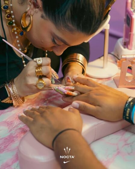 A woman wearing various wristbrands for different events is getting her nails done. The woman doing her nails wears lavishly long acrylics in Floridian sunset tones, some branded with the word NIKE or the brand's logo. She also wears a stack of gold bracelets and several chunky rings, one is a capital P.