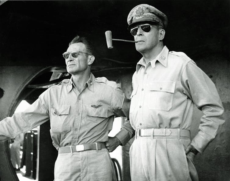 Two men in khakis, wearing sunglasses. MacArthur smokes a pipe.