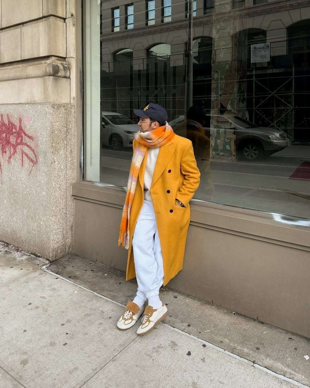 stylish man wearing a mustardy yellow topcoat, orange and lavender scarf, navy baseball hat, and off-white sweatshirt and joggers with white and tan sneakers