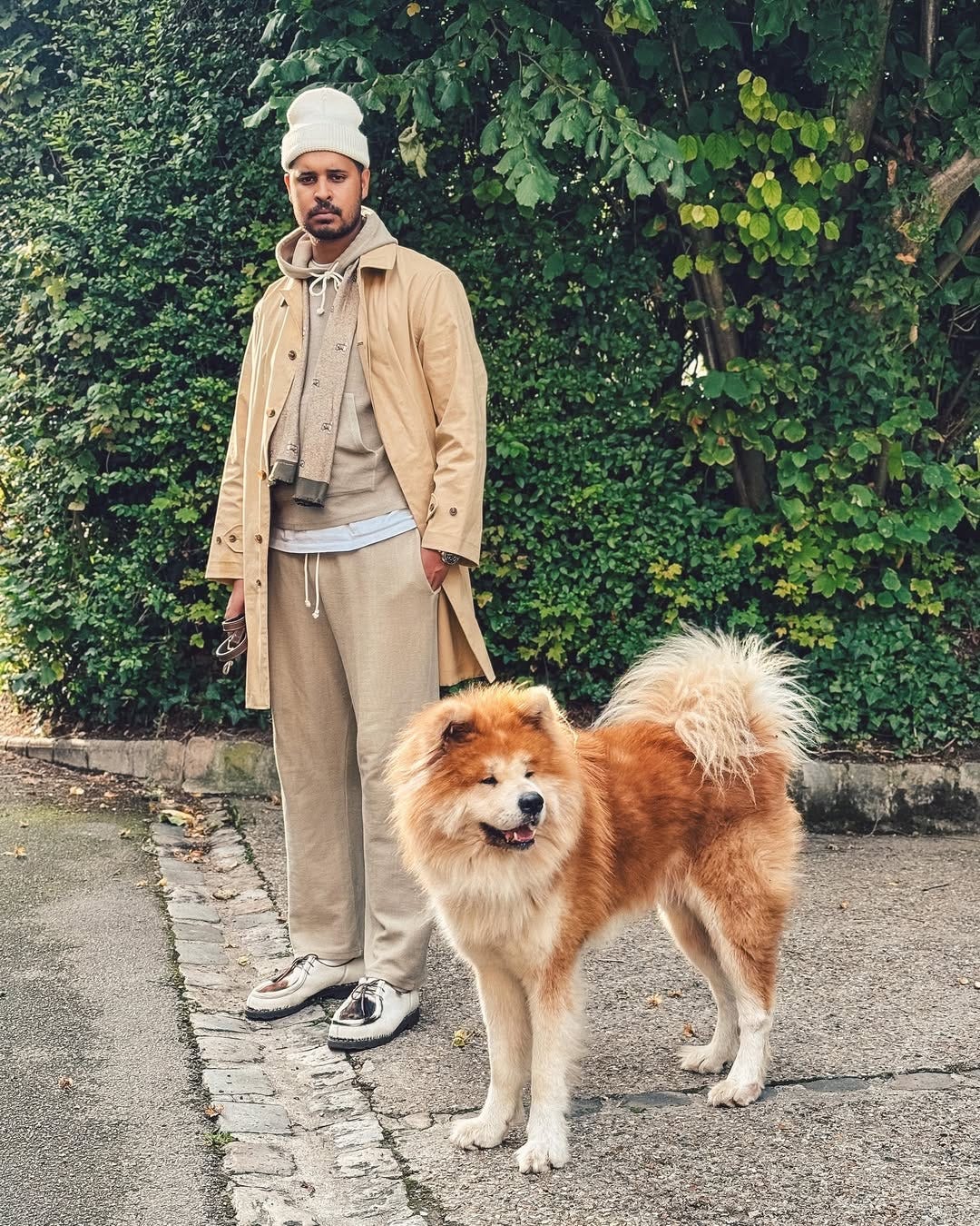 stylish man wearing tan drawstring pants with a tan hoodie and tan topcoat, standing outside next to a fluffy dog