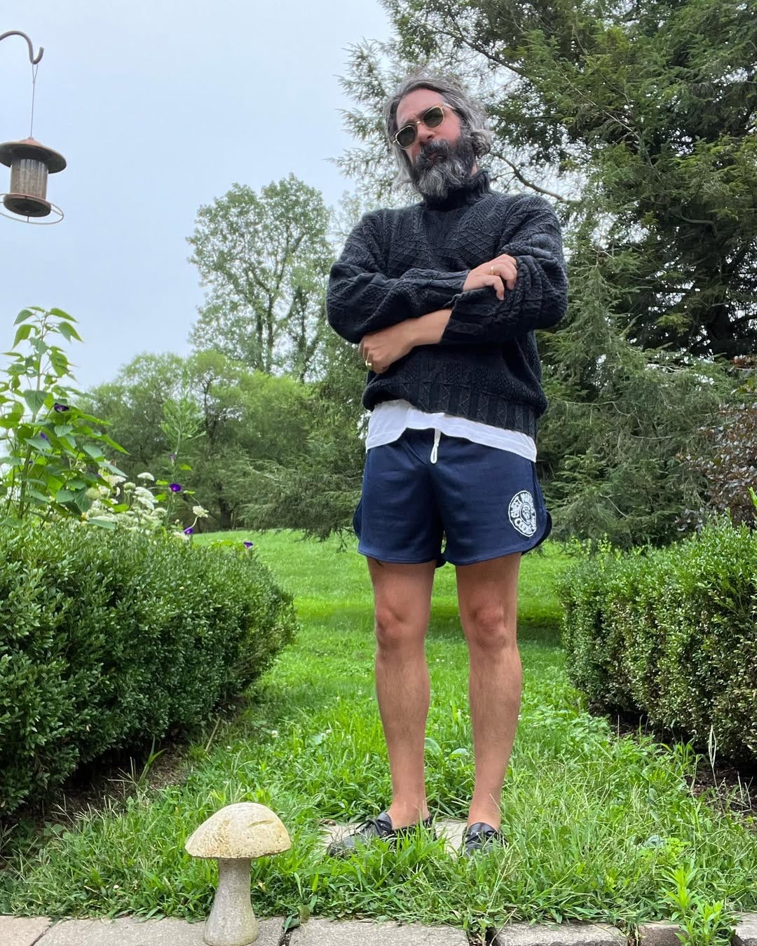 stylish bearded man wearing a textured grey turtleneck over a white t-shirt with blue athletic shorts and brown boat shoes