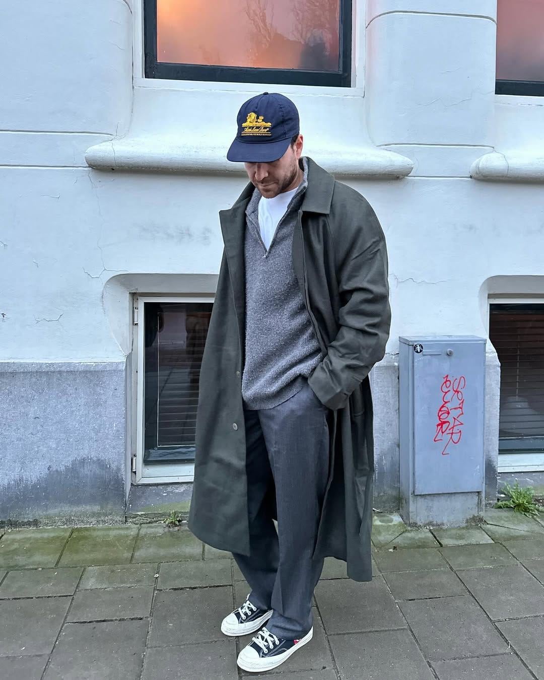 stylish young man wearing a grey quarter zip sweater over a white t-shirt with grey pants and a dark topcoat with sneakers