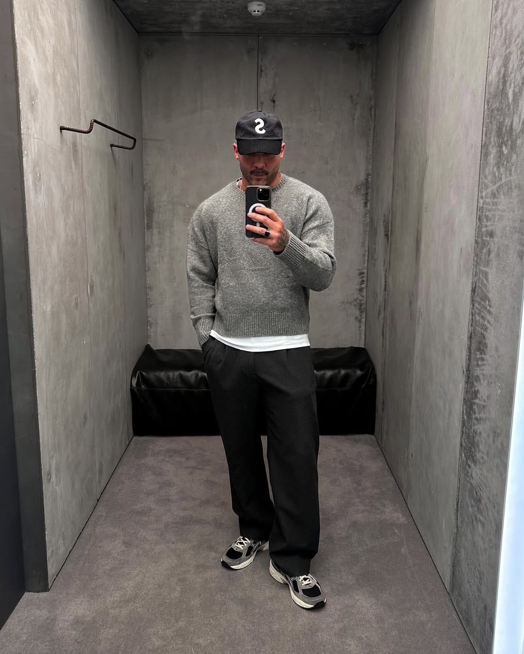 stylish man wearing a grey crewneck sweater, white t-shirt, and black pants taking a selfie in a dressing room mirror