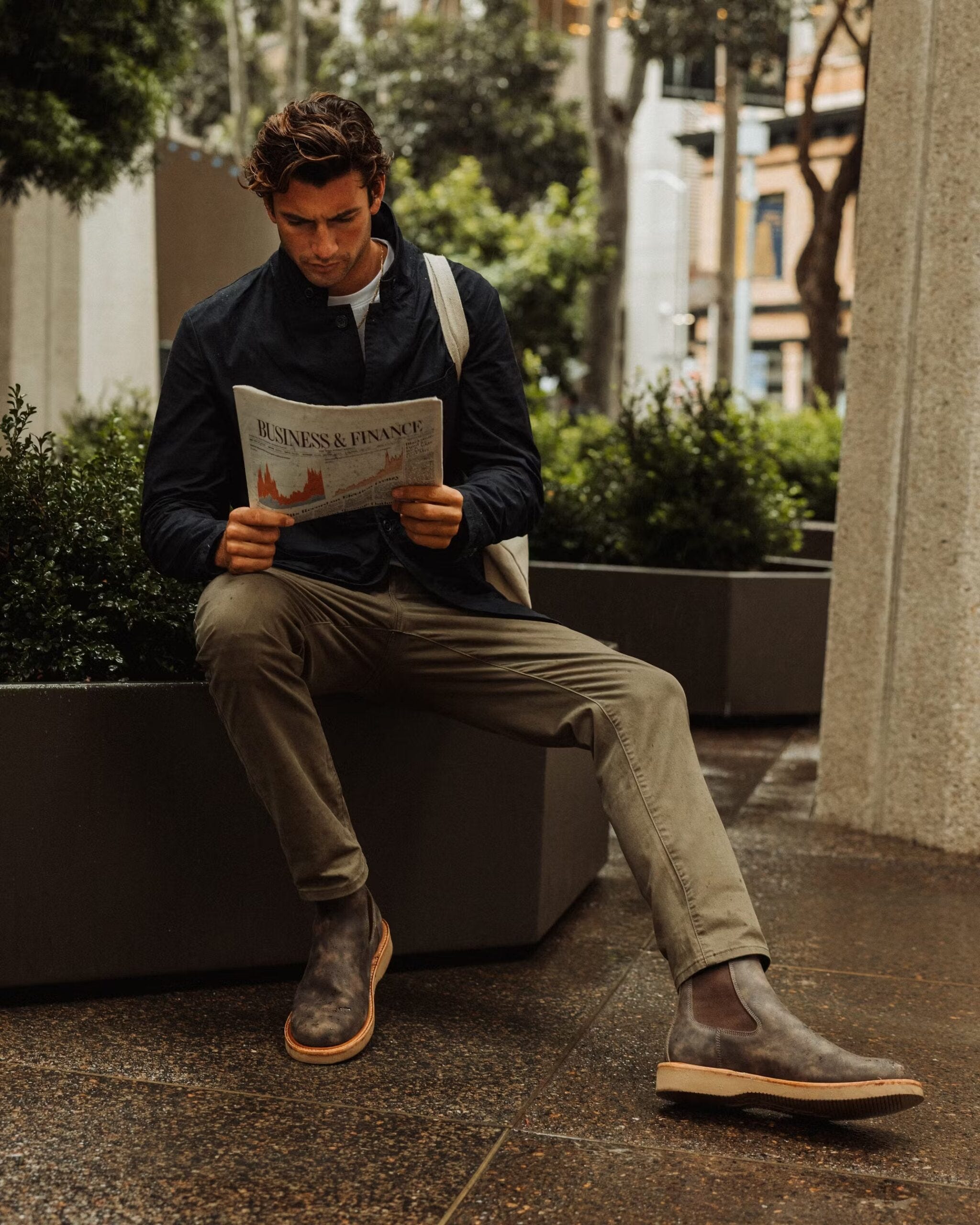 stylish man sitting down reading a newspaper and wearing a navy jacket and tan pants with brown boots