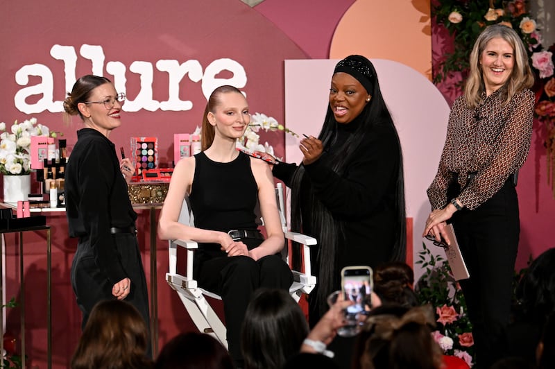 In front of the Allure magazine logo and a table of Pat McGrath Labs products, McGrath demonstrates a soft, rosy glam makeup look on the face of a model with sleek pulled-back hair.